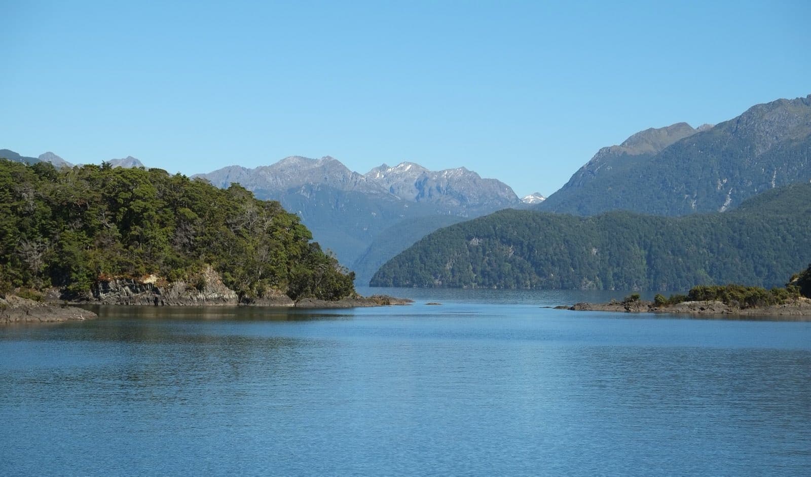 Calm blue water and forested islands in Lake Manapouri with mountains beyond